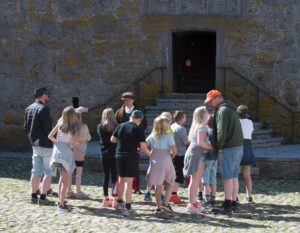 Guide i medeltidskläder och skolklass med lärare samlade framför borgen ingång.