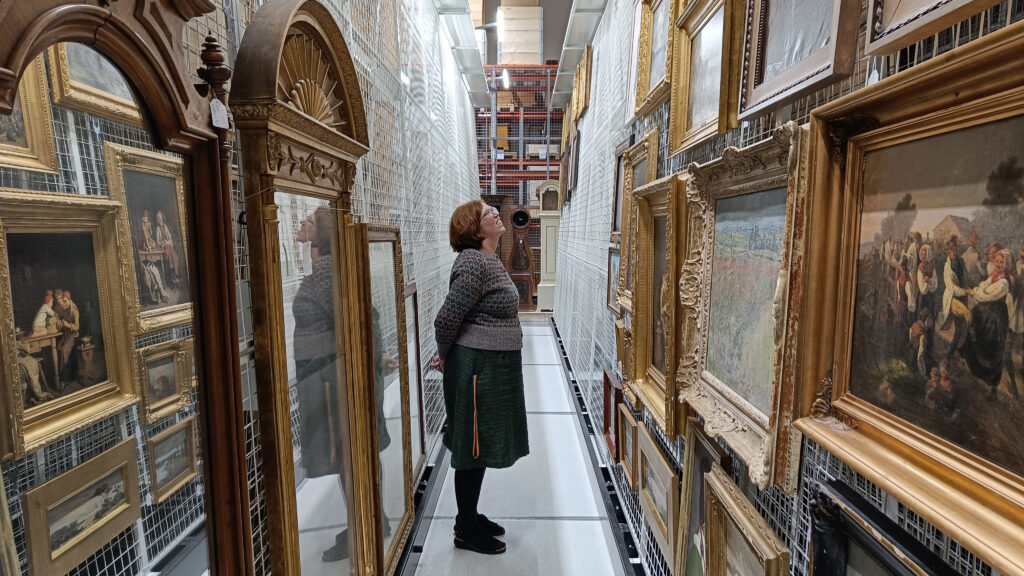 En kvinna står i ett magasin och tittar uppåt väggen; i förgrunden hänger målningar.