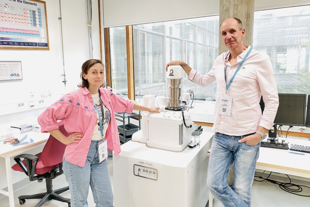 En kvinna i rosa jacka och en man i rosa skjorta står vid en apparat i ett laboratorium.