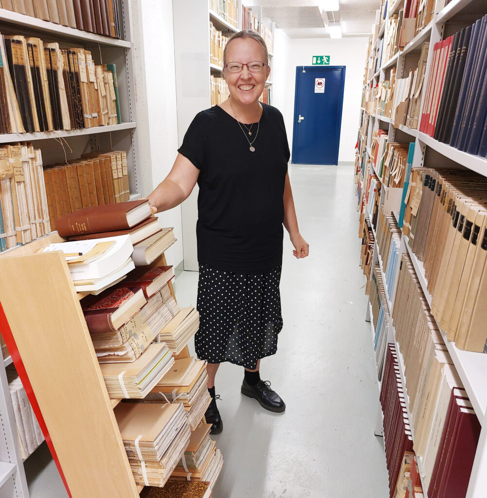 En kvinna mellan bokhyllor i ett arkiv. Hon ler.
