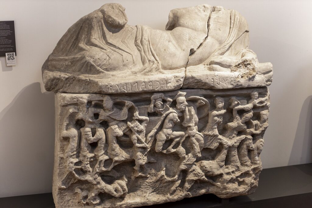 En gravurna i sten med många uthuggna detaljer av människofigurer.