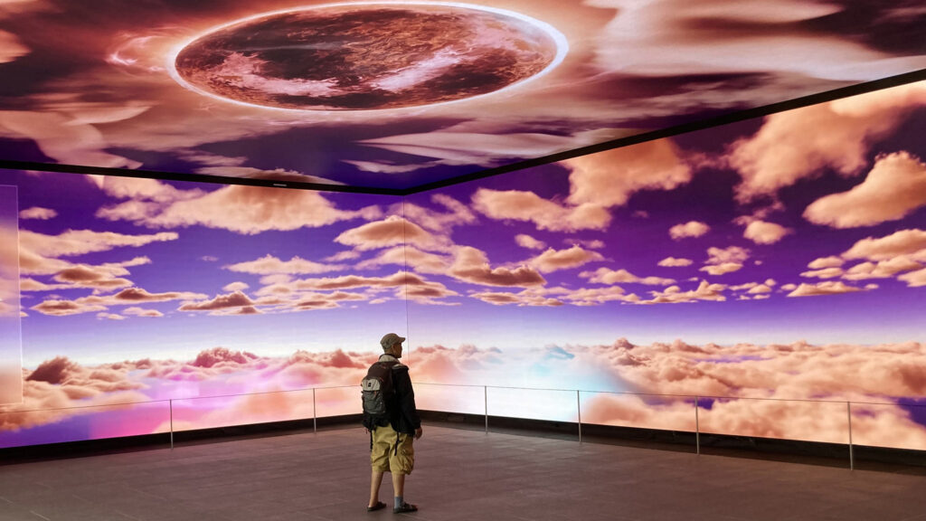 En person står inuti en utställningssal med projektioner på väggar och tak.