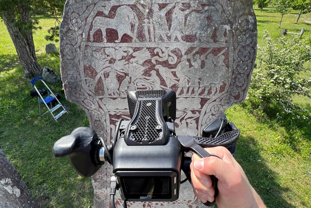 En hand håller en kamera framför en bildsten med olika figurer på.