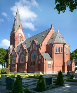 Stor kyrka i rött tegel med grått tak. Gravstenar i förgrunden.