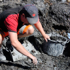 Arkeolog som sitter på huk och gräver i Motala. Han har keps och håller i ett spann och en kratta.