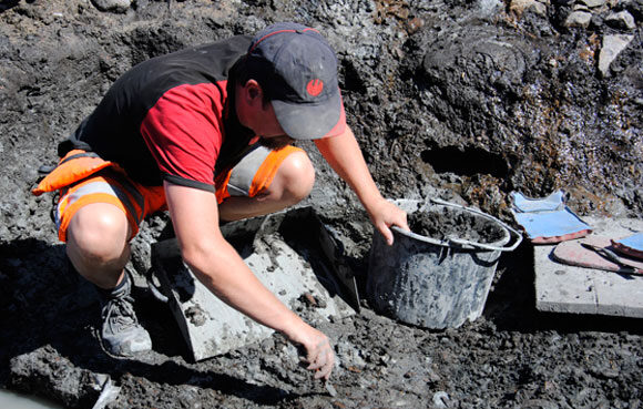 Arkeolog som sitter på huk och gräver i Motala. Han har keps och håller i ett spann och en kratta.