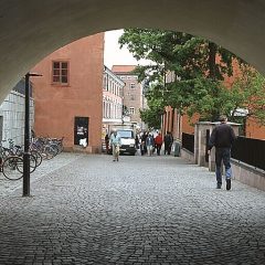 Gatuvy i Uppsala, med en stensatt gata som går igenom ett valv, och cyklar, promenerande människor och en bil ses i bakgrunden