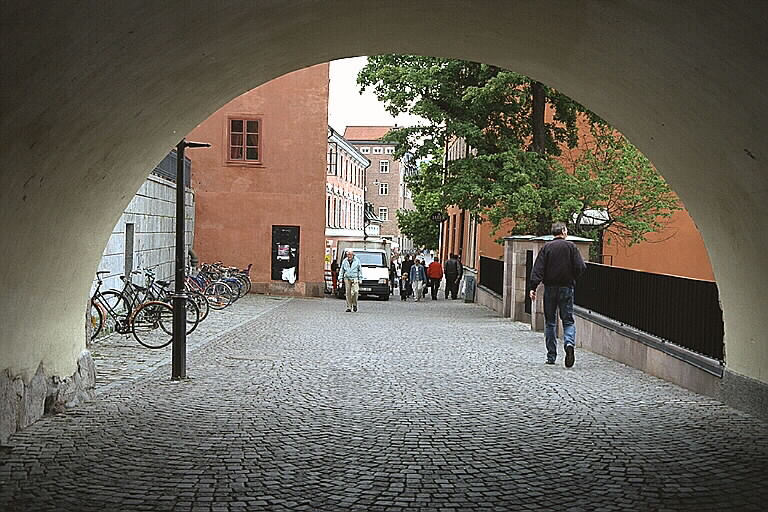 Gatuvy i Uppsala, med en stensatt gata som går igenom ett valv, och cyklar, promenerande människor och en bil ses i bakgrunden