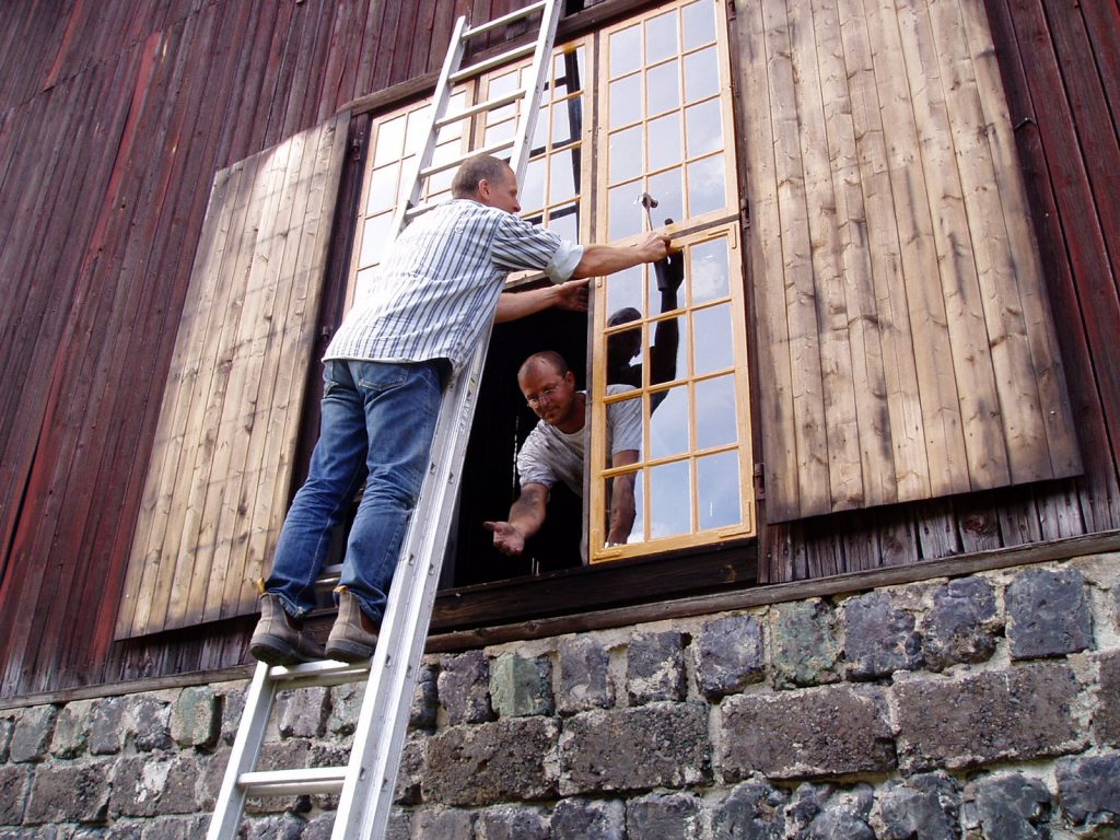 Två personer renoverar ett fönster. En står på en stege och den andra tittar ut och handräcker.