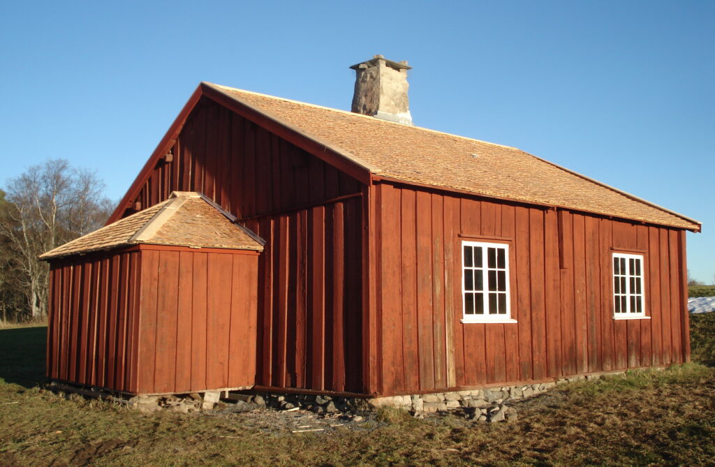 En räd träbyggnand med spåntak. På långsidan två vitmålade spröjsfönster och på kortsidan en rund utbyggnad.