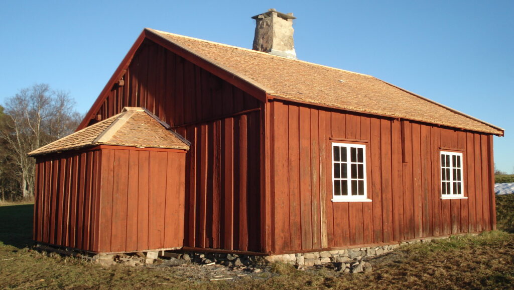 En räd träbyggnand med spåntak. På långsidan två vitmålade spröjsfönster och på kortsidan en rund utbyggnad.