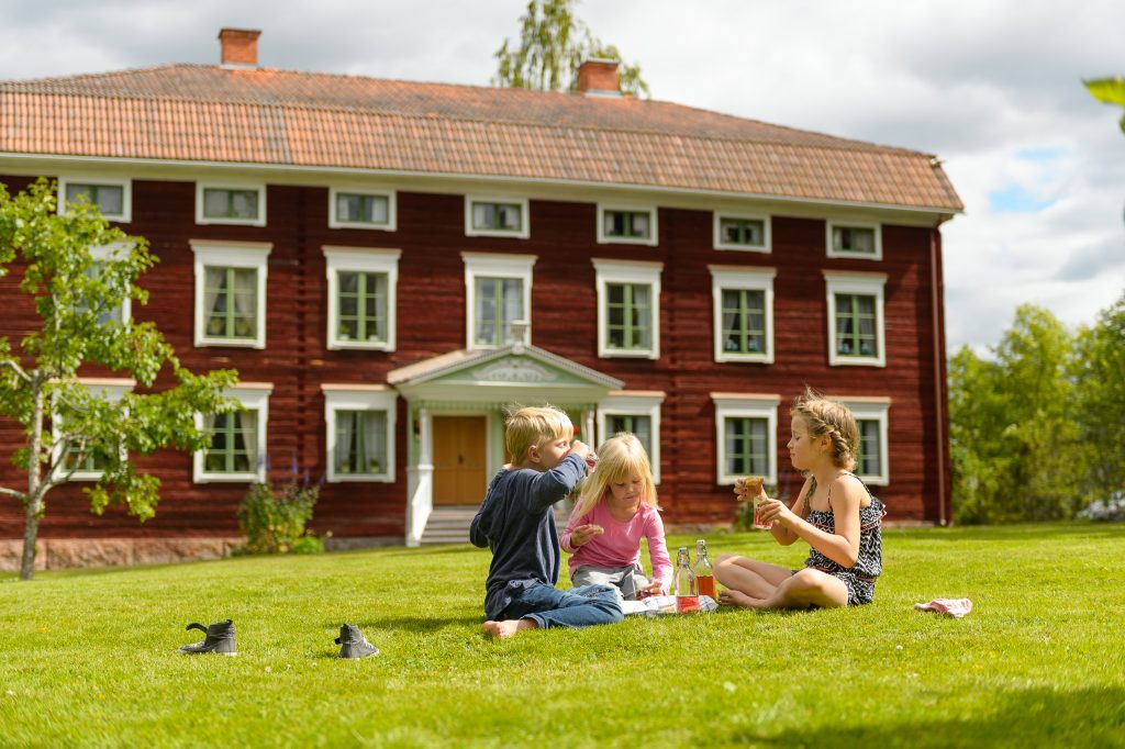 Några barn sitter på gräsmattan vid en stor rödmålad äldre byggnad med en elegant dekorerad brokvist. Huset är byggt i trä och har tre synliga våningar med fönster med vita karmar.