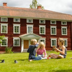 Några barn sitter på gräsmattan vid en stor rödmålad äldre byggnad med en elegant dekorerad brokvist. Huset är byggt i trä och har tre synliga våningar med fönster med vita karmar.