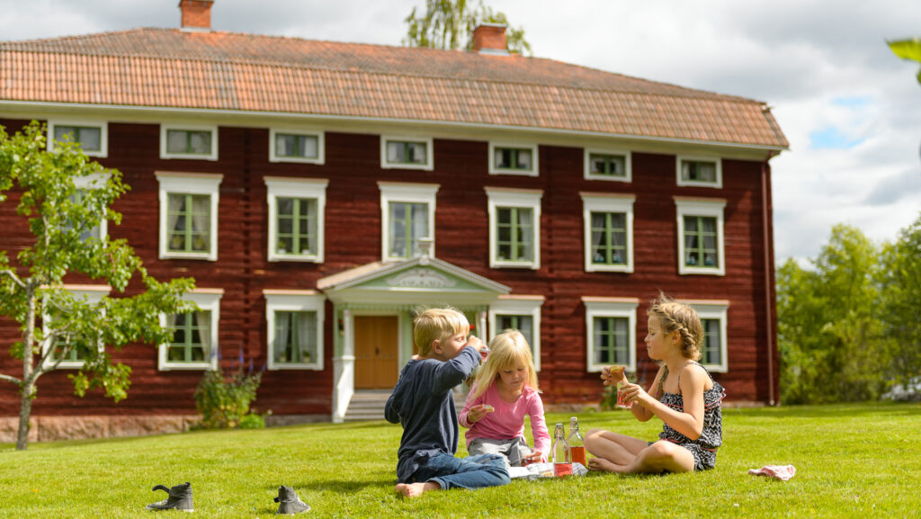 Några barn sitter på gräsmattan vid en stor rödmålad äldre byggnad med en elegant dekorerad brokvist. Huset är byggt i trä och har tre synliga våningar med fönster med vita karmar.