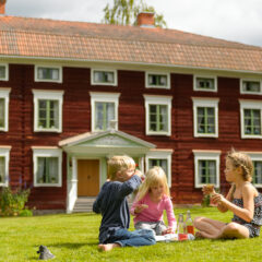 Några barn sitter på gräsmattan vid en stor rödmålad äldre byggnad med en elegant dekorerad brokvist. Huset är byggt i trä och har tre synliga våningar med fönster med vita karmar.