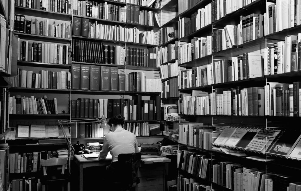 En svartvit bild på ett litet rum med väggarna täckta av böcker. En kvinna sitter vid ett skrivbord med ryggen mot kameran.