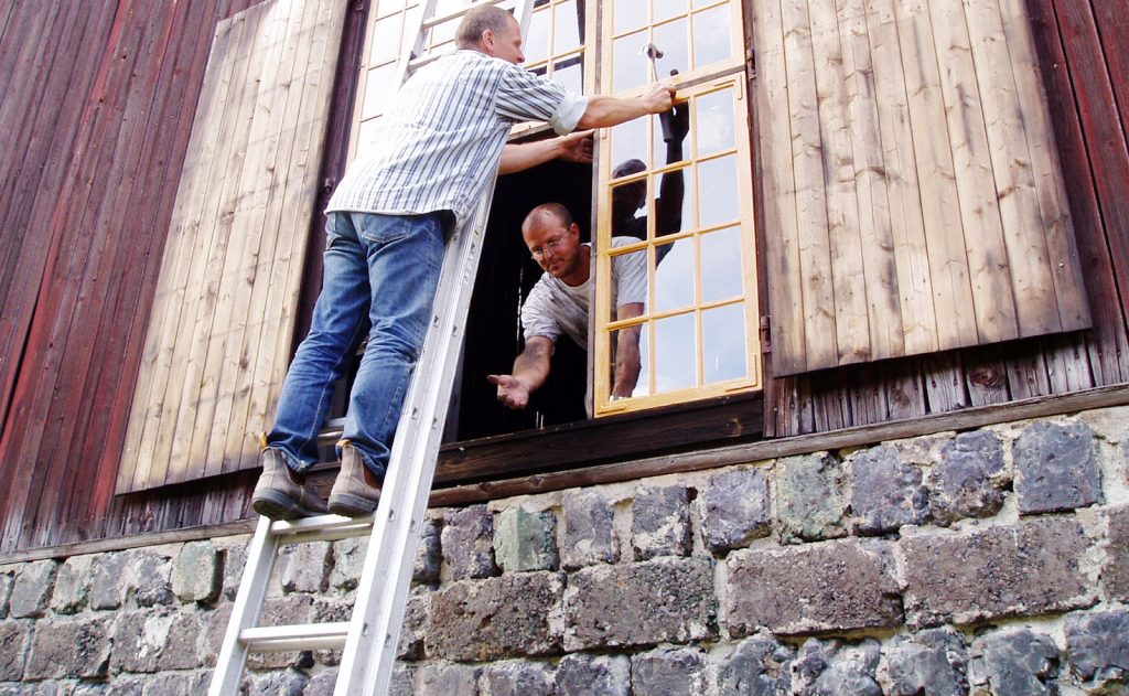 Två personer renoverar ett fönster. En står på en stege och den andra tittar ut och handräcker.