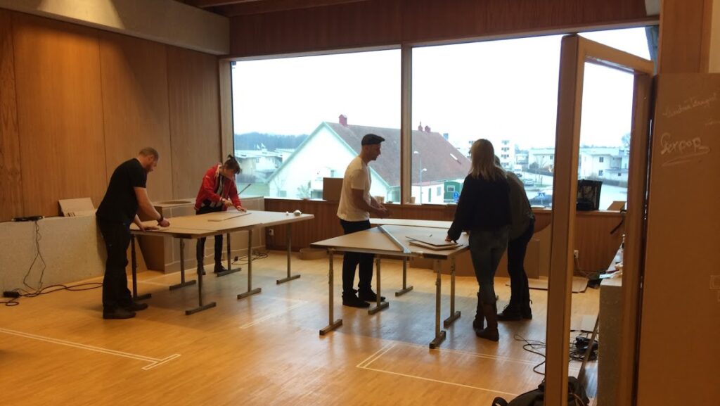 Fem personer står lutade över arbetsbord och utför olika moment.