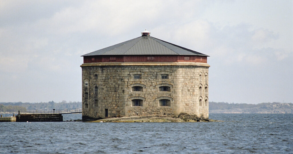 Gult kantigt tegeltorn i fyra våningar med fönstergluggar åt varje håll, ligger på ett litet skär mitt i havet.