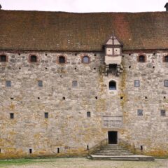 Borgens högra gråa fasad med små gluggar, en trappgavel och rött tegeltak, bredvid skymtar ett gult trevångingshus som är hälften så högt.