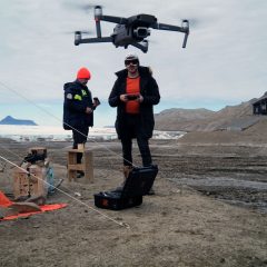 Två män står utanför ett orange tält i ett kargt bergslandskap. En av männen flyger en drönare.