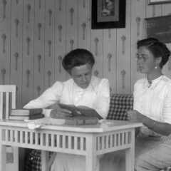 Svart-vit historisk bild (1911) med två kvinnor klädda i vitt, sittande vid ett bord, båda läsande – inomhusmiljö med bord och möbler.