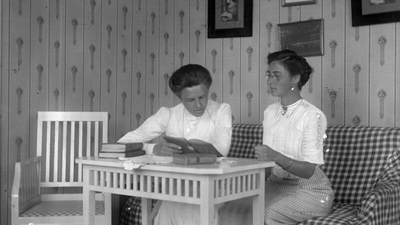 Svart-vit historisk bild (1911) med två kvinnor klädda i vitt, sittande vid ett bord, båda läsande – inomhusmiljö med bord och möbler.