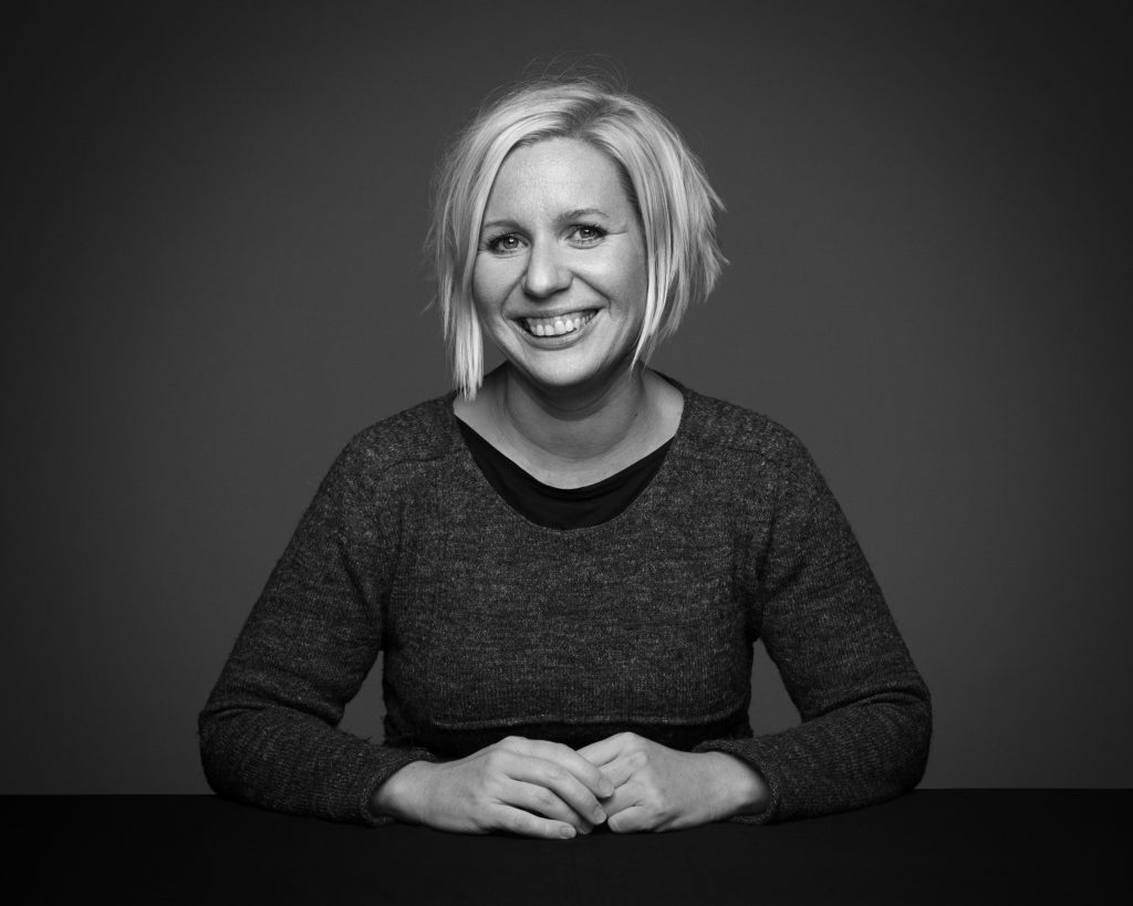 Ett svartvitt fotografi på Karin Nilsson. hon har mörka kläder på sig och ler stort.