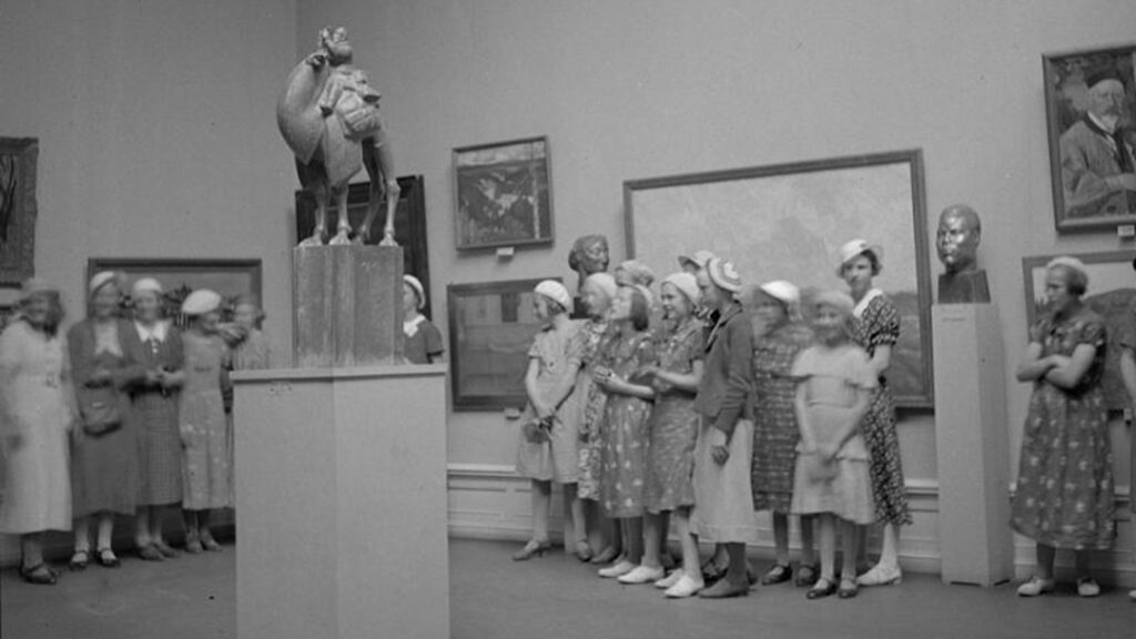 Svartvit bild med en grupp damer alla iklädda basker och lång kjol står framför ett musieföremål.