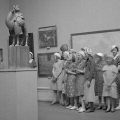 Svartvit bild med en grupp damer alla iklädda basker och lång kjol står framför ett musieföremål.