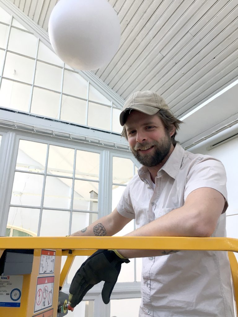 Simon deBrée utställningstekniker på Ronneby konsthall. Står på en gul lift i vit skjorta och keps.