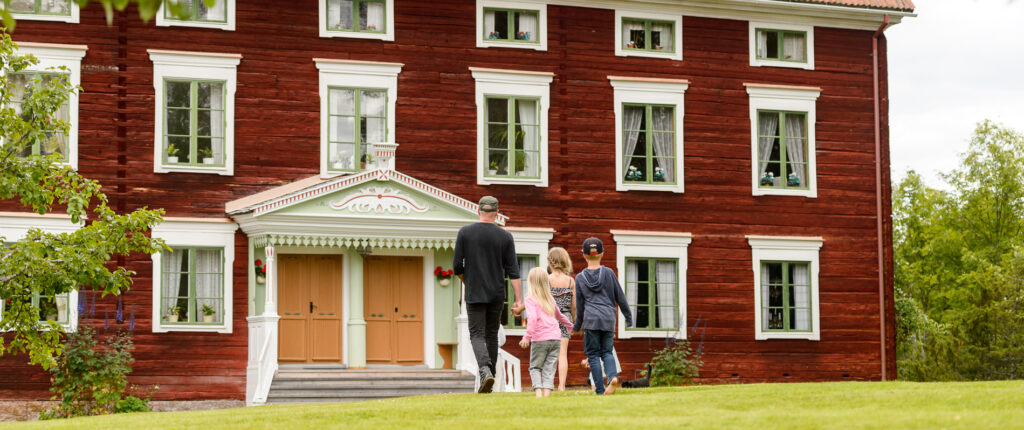 Några personer går över gräsmattan mot en stor rödmålad äldre byggnad med en elegant dekorerad brokvist. Huset är byggt i trä och har tre synliga våningar med fönster med vita karmar.