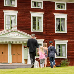 Några personer går över gräsmattan mot en stor rödmålad äldre byggnad med en elegant dekorerad brokvist. Huset är byggt i trä och har tre synliga våningar med fönster med vita karmar.