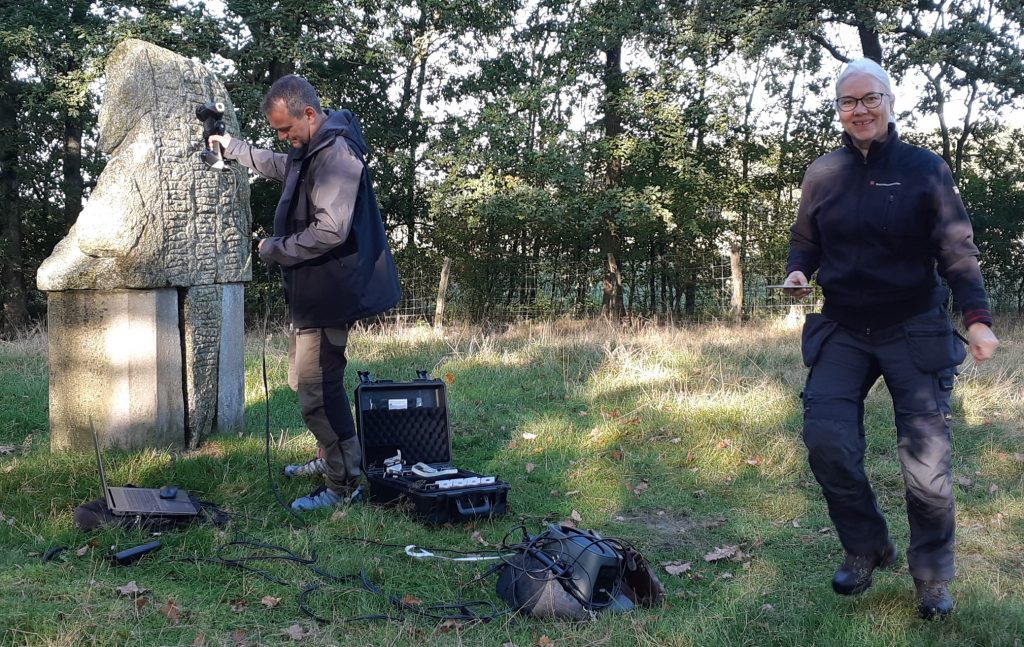 Till höger en kvinna som går mot kameran. Hon är klädd i arbetsbyxor och arbetsskor. Till vänster en man vid en stor runsten. Han håller en apparat mot runstenen. På marken ligger väskor med datautrustning. Gräset är gult och det ligger torra höstlöv på marken.