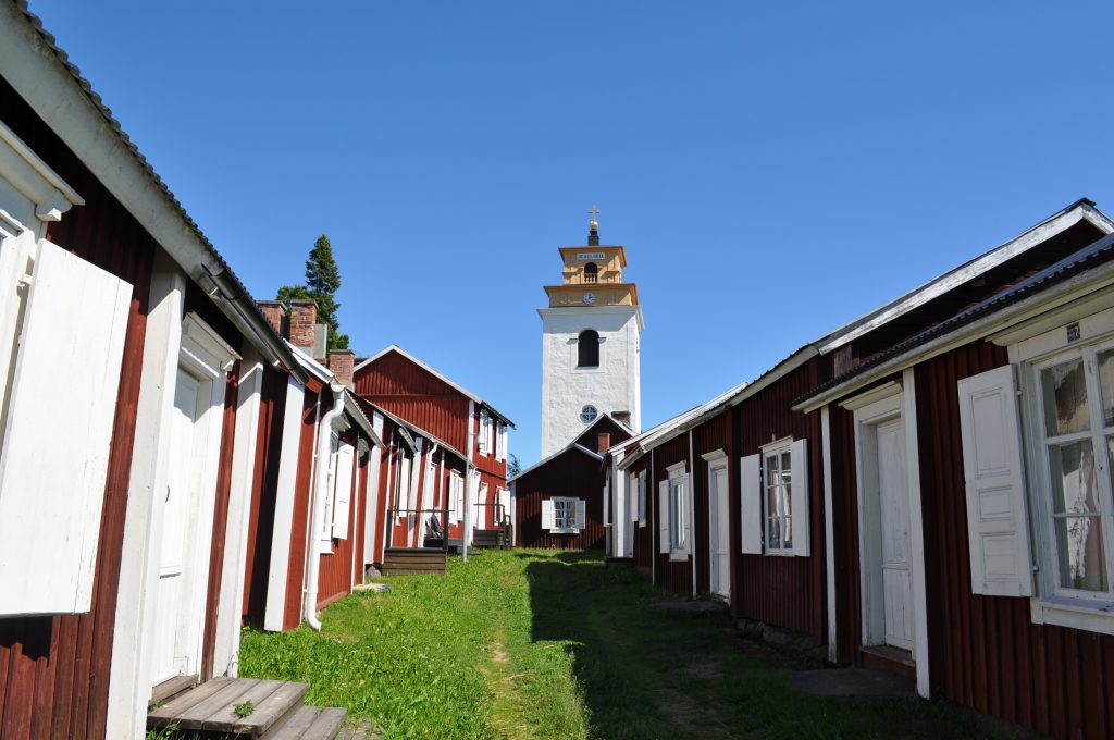 En lång rad med röda träbyggnader med vita knutar, mellan dem en grönskanda stig som leder fram till en kyrka som ståtligt tornar mot blå himmel.