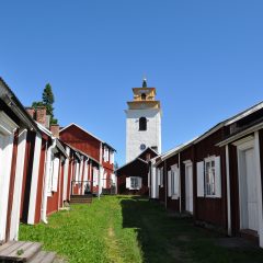 En lång rad med röda träbyggnader med vita knutar, mellan dem en grönskanda stig som leder fram till en kyrka som ståtligt tornar mot blå himmel.