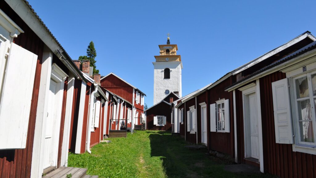 En lång rad med röda träbyggnader med vita knutar, mellan dem en grönskanda stig som leder fram till en kyrka som ståtligt tornar mot blå himmel.