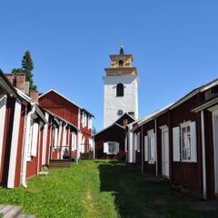 En lång rad med röda träbyggnader med vita knutar, mellan dem en grönskanda stig som leder fram till en kyrka som ståtligt tornar mot blå himmel.