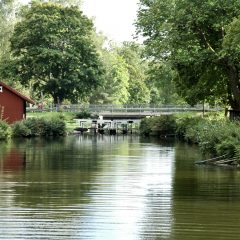 Vy över Kanalmuseet Skantzen med kanalen i förgrunden där grönskan speglar sig i vattnet och i bakgrunden en bro över slussen.