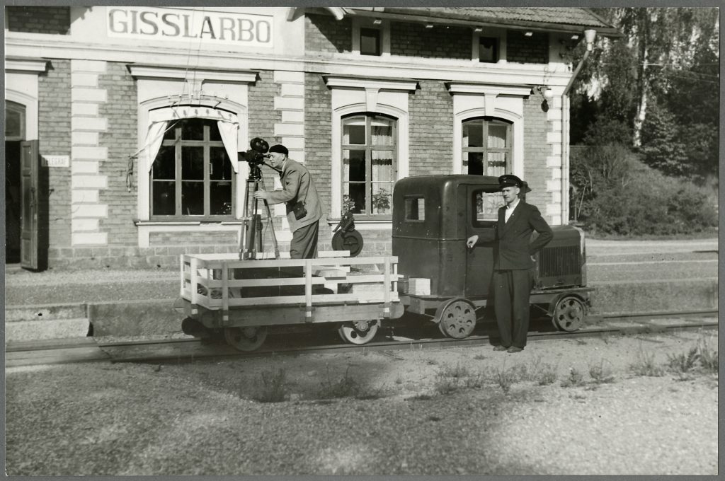 En man med basker står med filmkamera på en motordressin. Vagnen står på rälsen framför ett stationshus. Bredvid står en annan man