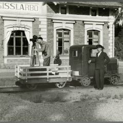 En man med basker står med filmkamera på en motordressin. Vagnen står på rälsen framför ett stationshus. Bredvid står en annan man