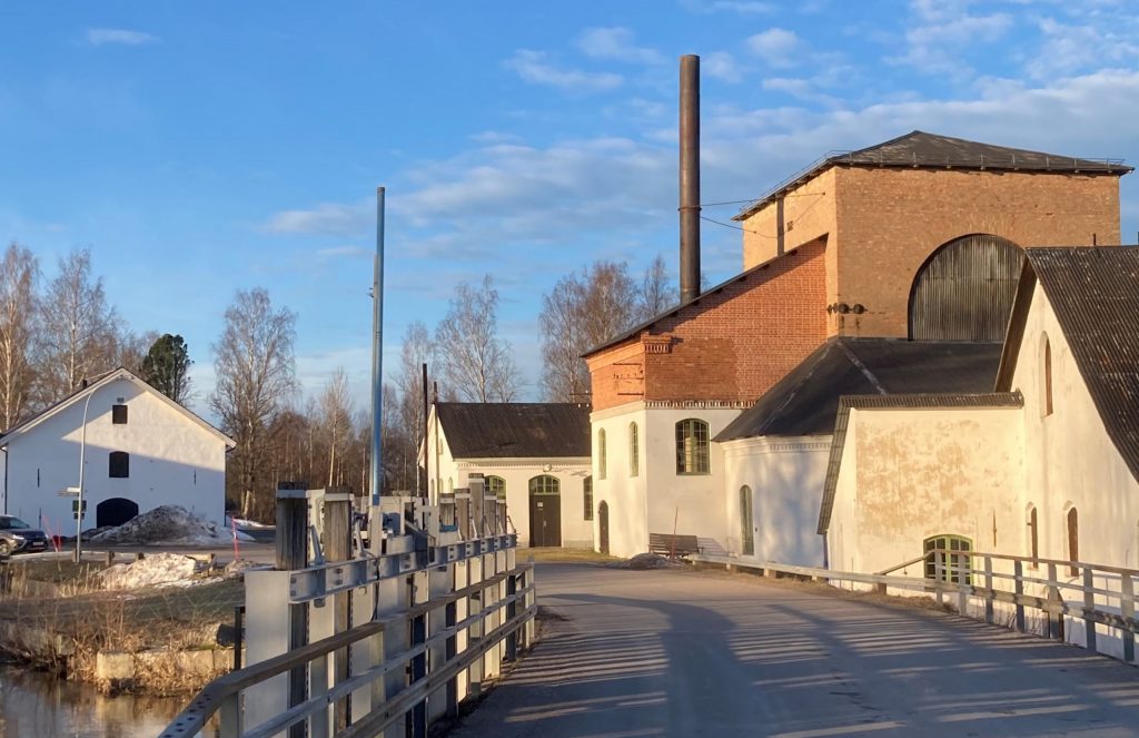 Vy över tegelbyggnad i bruksområde. I förgrunden en bro över vatten.