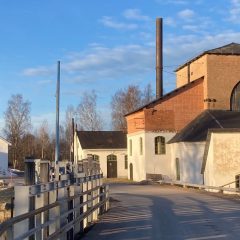 Vy över tegelbyggnad i bruksområde. I förgrunden en bro över vatten.