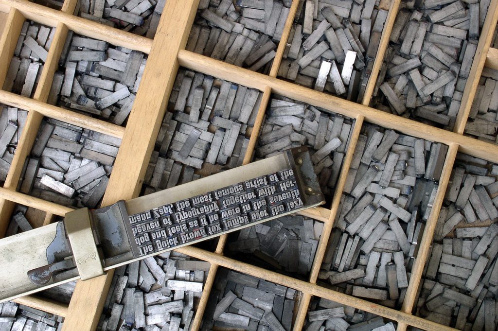 Närbild på massor av bokstavstyper i grå metall som ligger i en trädlåda.