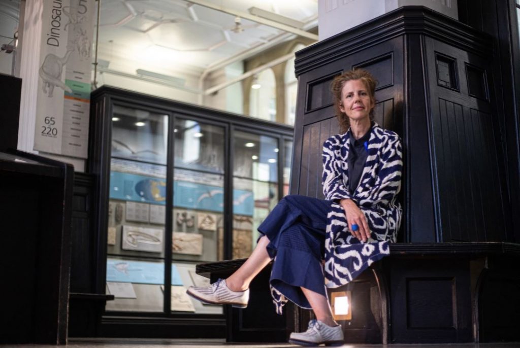 Manchester Museums direktör Esme Ward sitter framför en mörk träpanel i museibyggnaden och ser in i kameran.