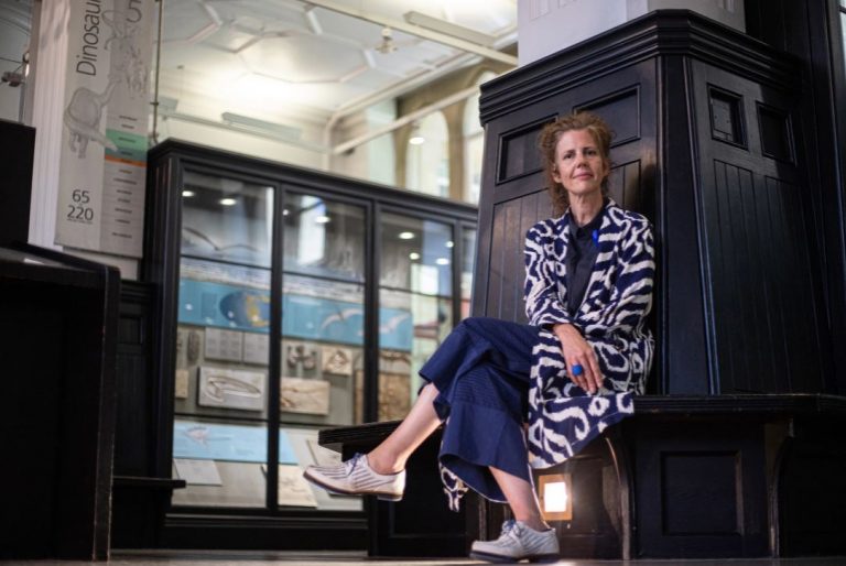 Manchester Museums direktör Esme Ward sitter framför en mörk träpanel i museibyggnaden och ser in i kameran.