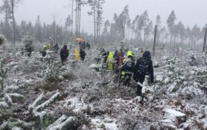 I ett snöoväder vandrar personer i ojämn skogsterräng med små granar.