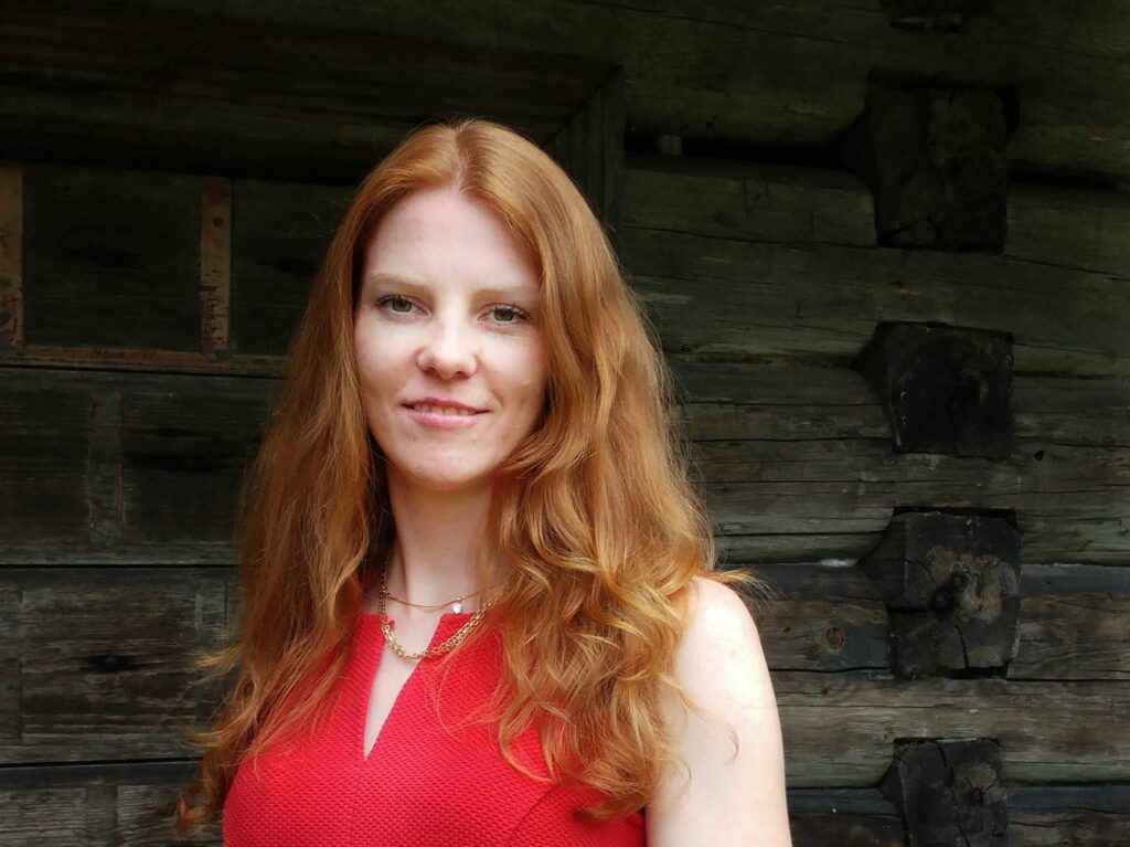 Porträttfotografi föreställande konservator Pia Edqvist iklädd en röd topp framför en timmervägg.