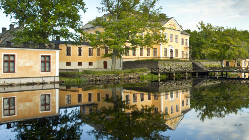 En gul herrgårdsbygnad i tre våningar speglas i en vattendamm.