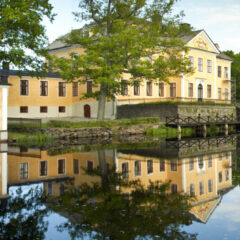 En gul herrgårdsbygnad i tre våningar speglas i en vattendamm.
